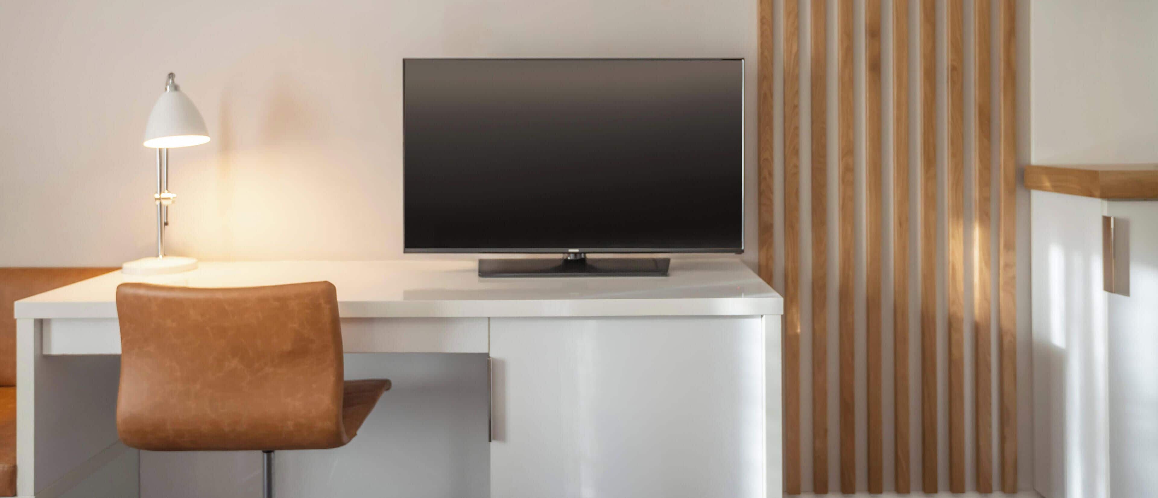 A modern room with a white desk, a flat-screen TV, a brown chair, and a lamp.