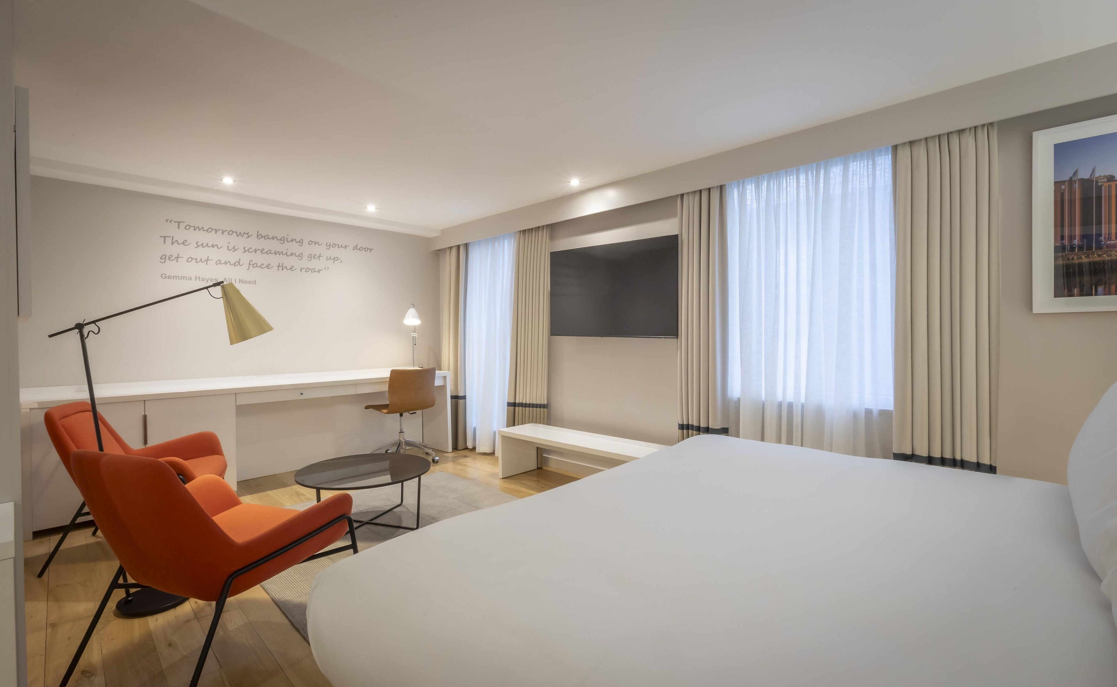 Ein Hotelzimmer mit Bett, Schreibtisch, Stuhl, Lampe, Fernseher und einem Fenster mit Vorhängen.