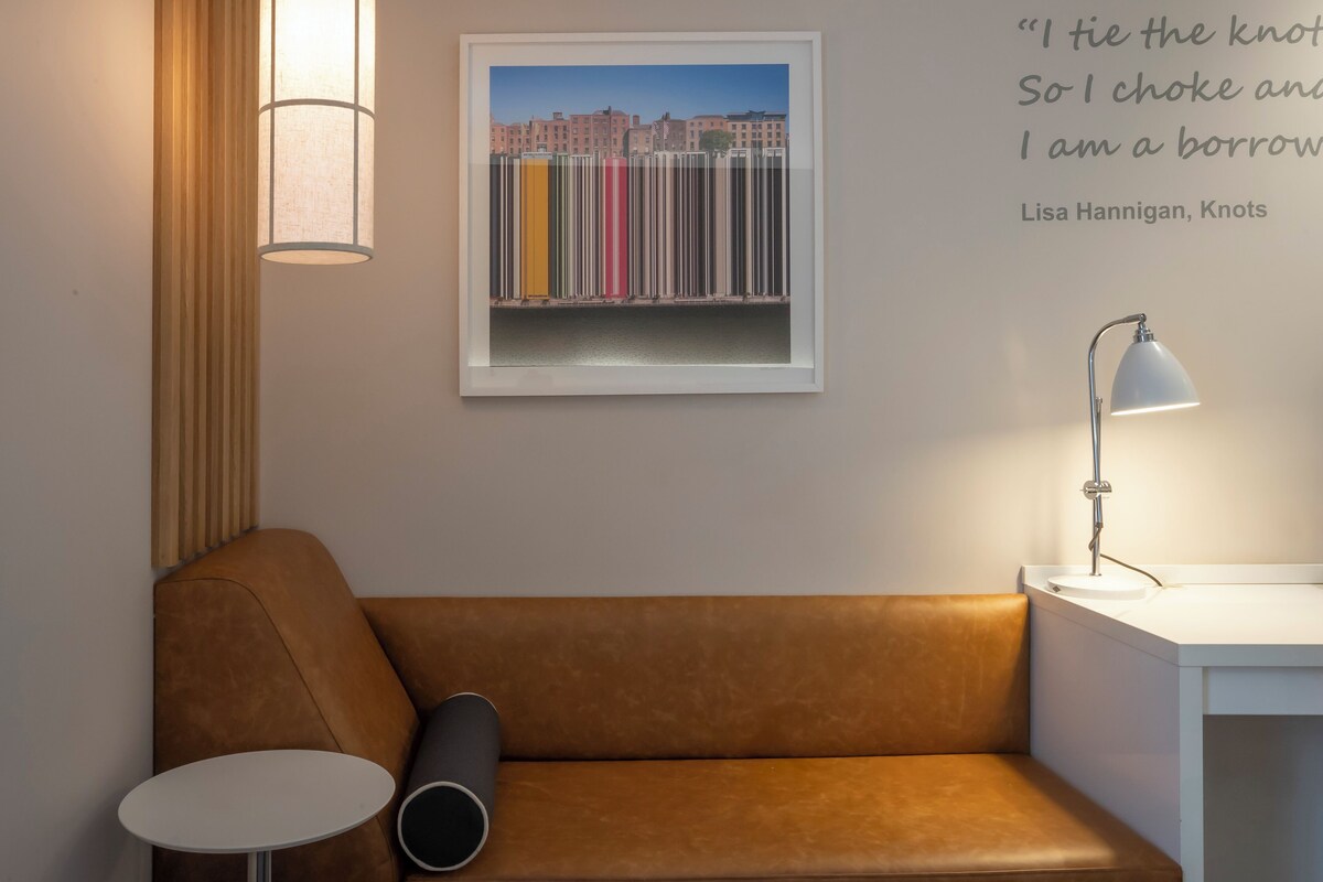 A modern room with a brown leather sofa, a small round table, a floor lamp, and a framed artwork of colorful buildings on the wall.