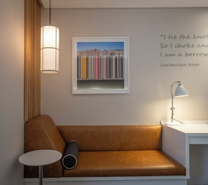 Una habitación moderna con un sofá de cuero marrón, una mesita redonda pequeña, una lámpara de pie y una obra de arte enmarcada con edificios coloridos en la pared.
