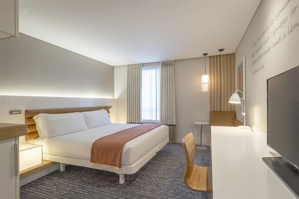 Quarto de hotel com cama, mesa de cabeceira, luminária, televisão e janela com cortinas.