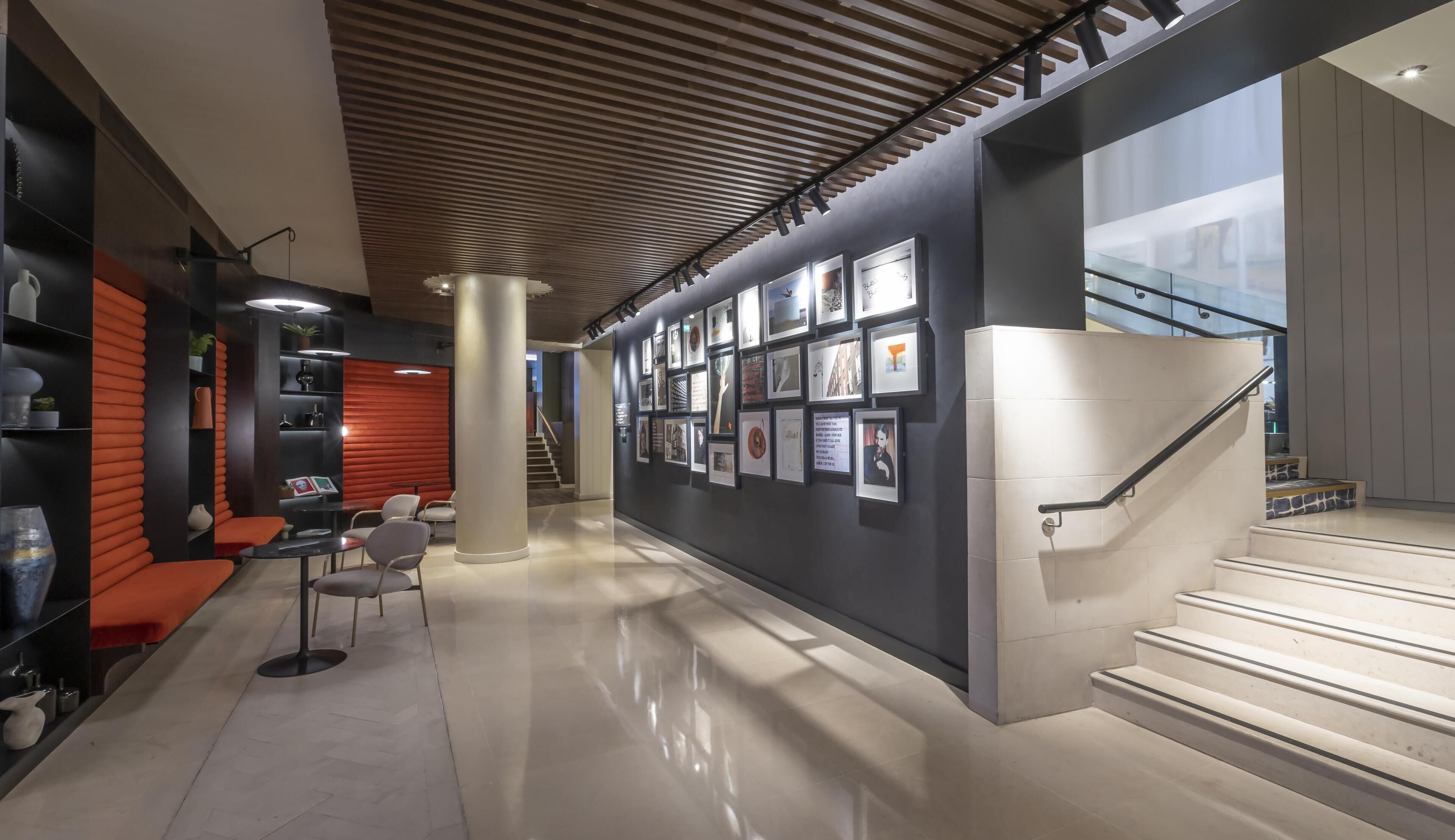 Un interior moderno con una escalera, una pared decorada con cuadros y una zona de estar con sillas y mesas.