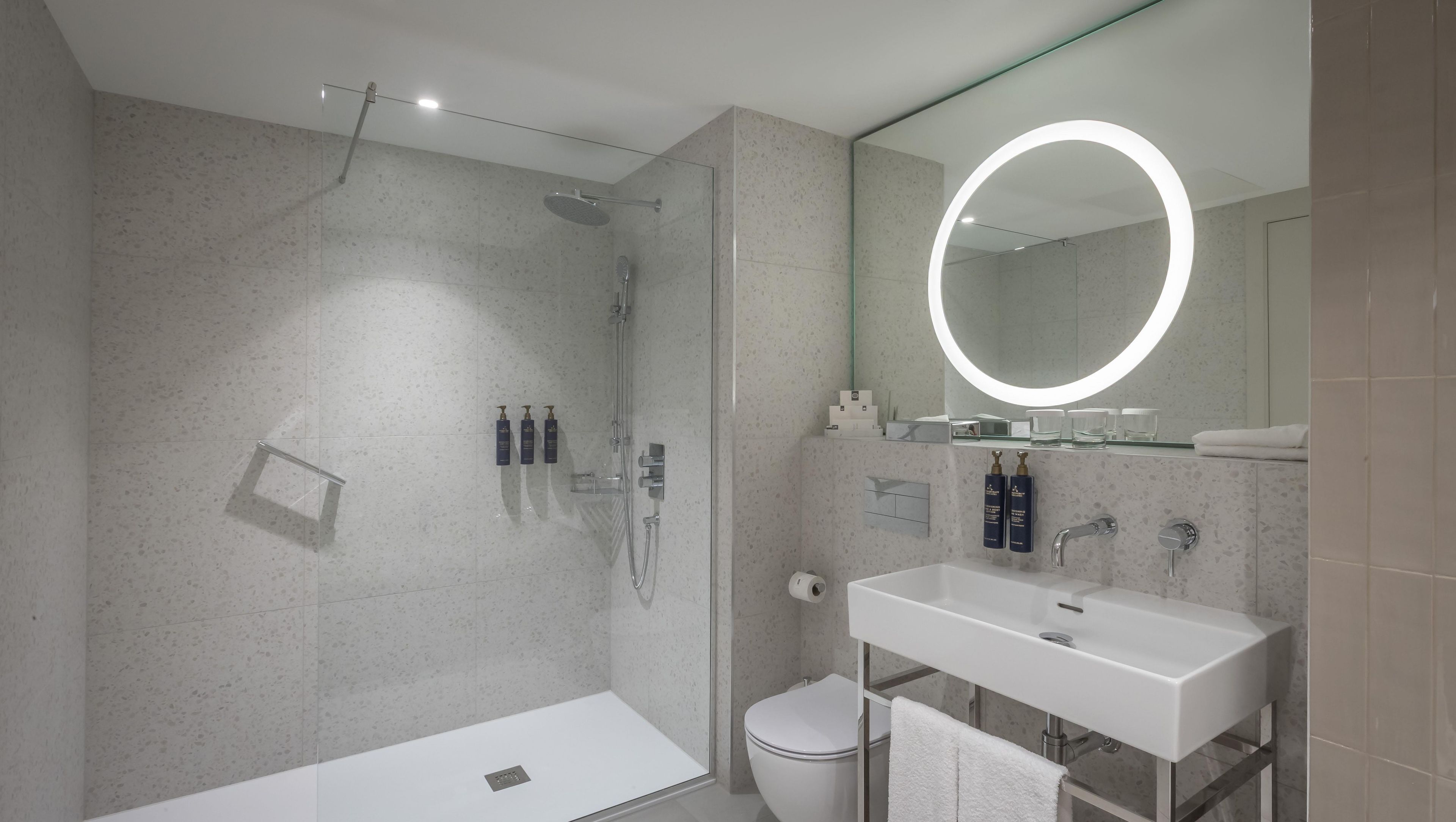 A modern bathroom with a glass-enclosed shower, a white sink, and a large mirror with a circular light.