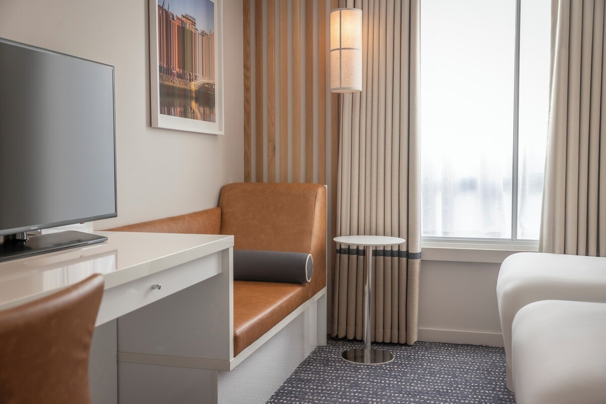 A hotel room with a desk, a brown leather chair, a white bed, a small round table, and a large window with curtains.