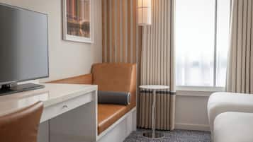 A hotel room with a desk, a brown leather chair, a white bed, a small round table, and a large window with curtains.