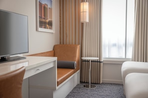 Una habitación de hotel con un escritorio, un sillón de cuero marrón, una cama blanca, una mesita redonda pequeña y un ventanal con cortinas.