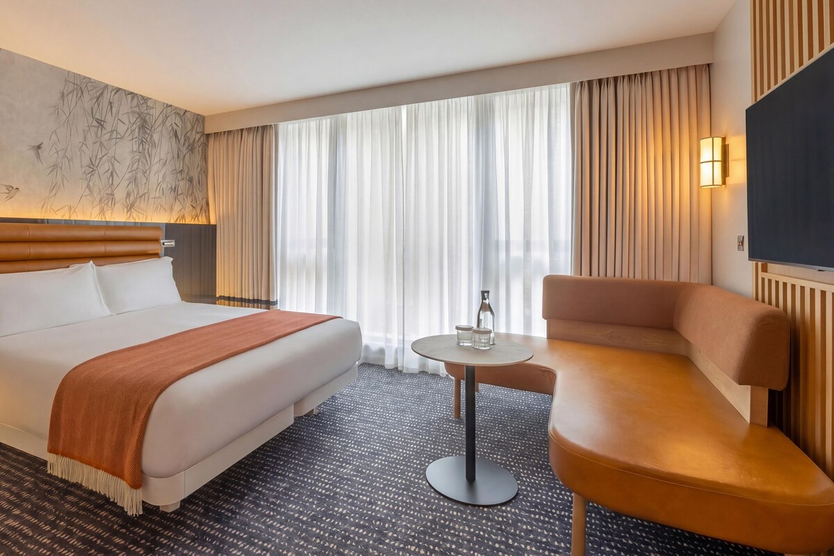 Ein Hotelzimmer mit einem großen Bett, einem kleinen runden Tisch, einer Couch, einem Fernseher und einem Fenster mit Vorhängen.
