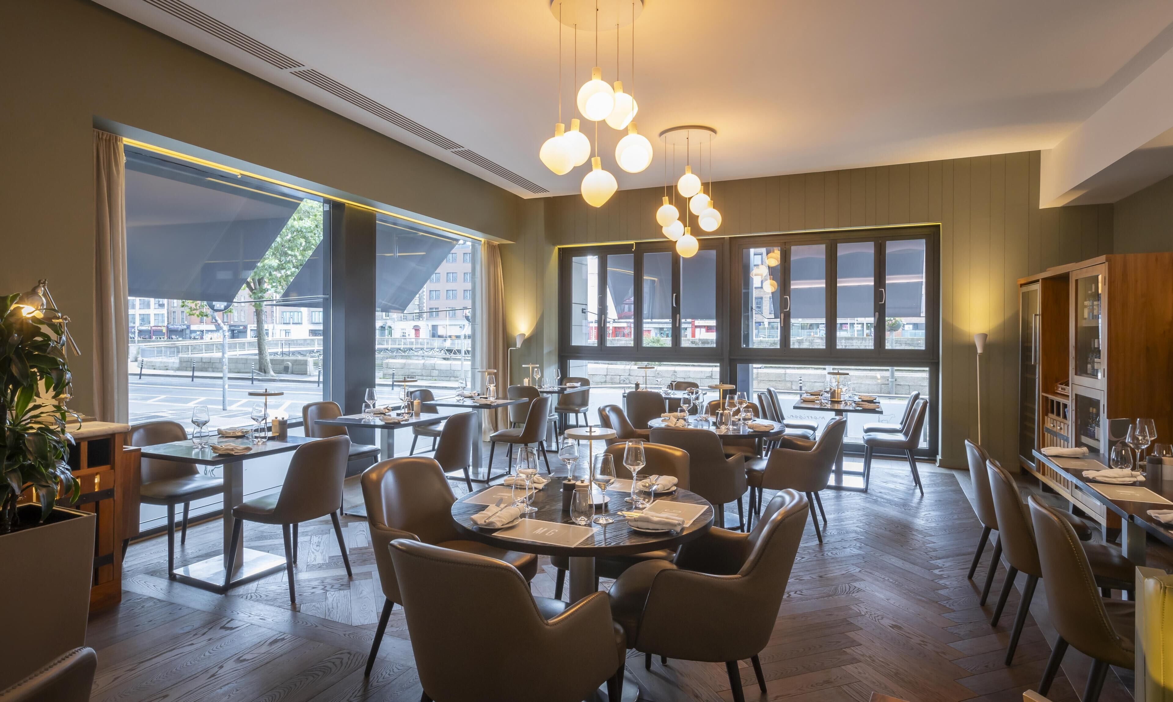 A restaurant with tables set for dining, large windows, and hanging pendant lights.