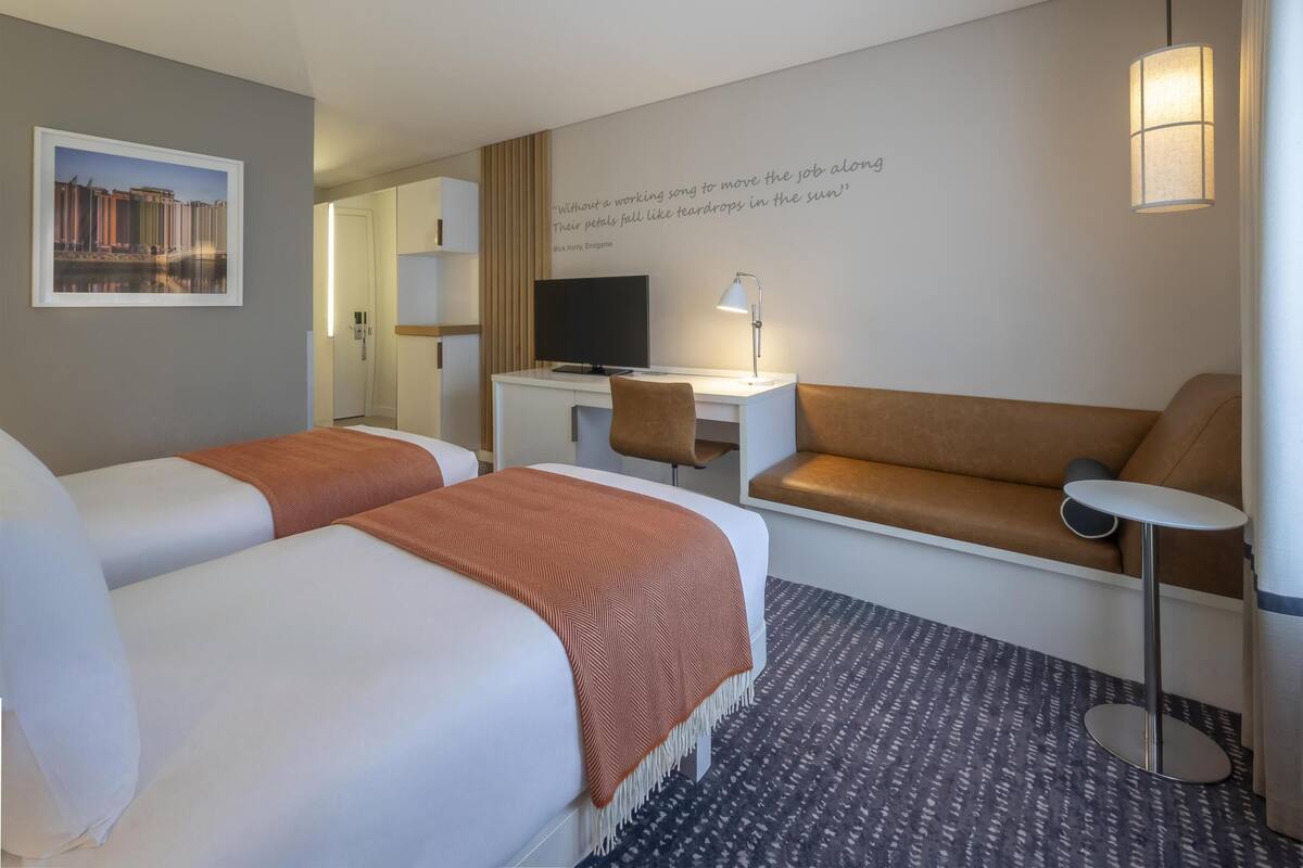 Quarto de hotel com duas camas, uma escrivaninha com computador, um sofá, uma mesinha e uma televisão presa à parede.