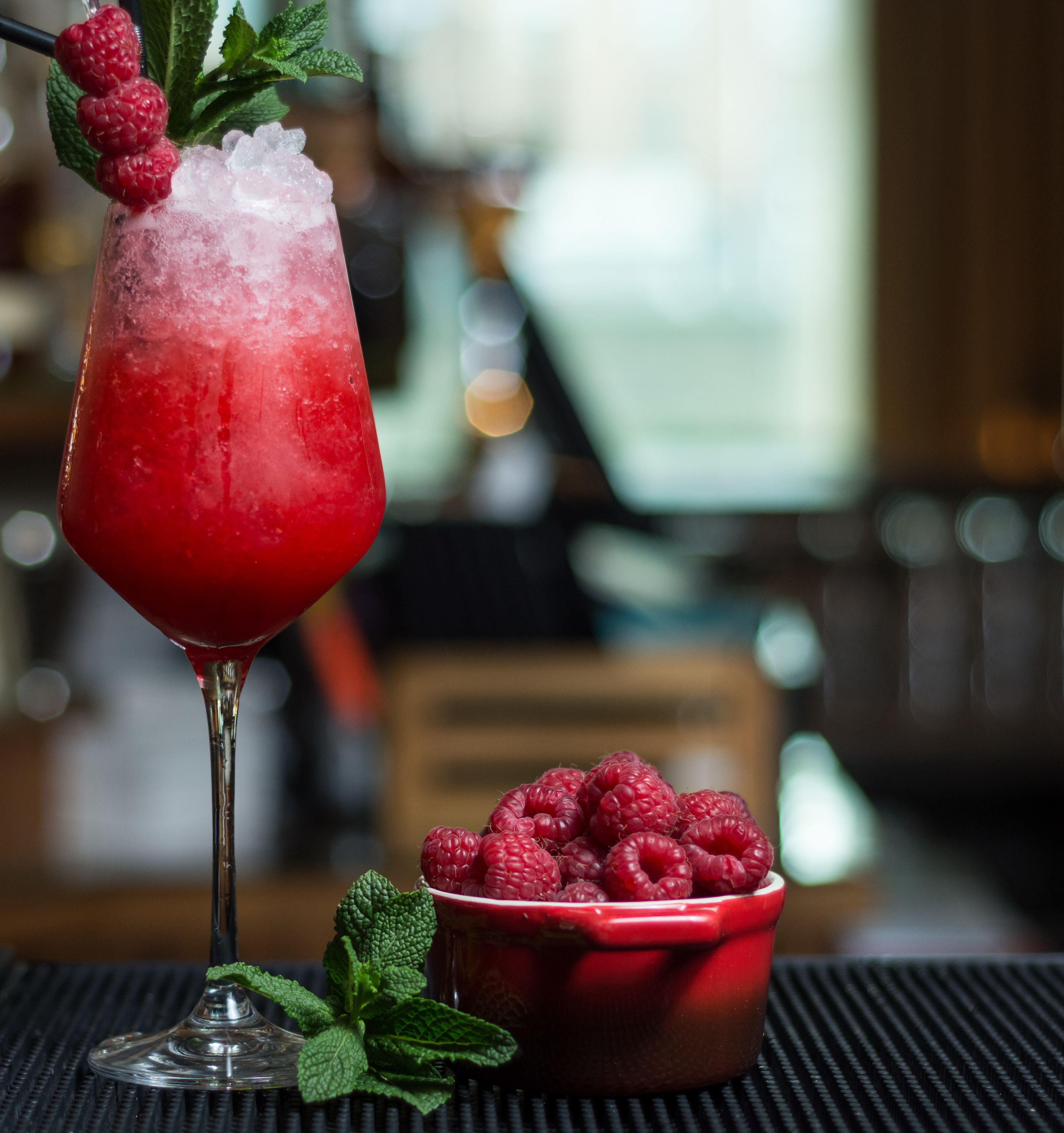 A cocktail glass with a red drink, ice, and raspberries on a black surface.