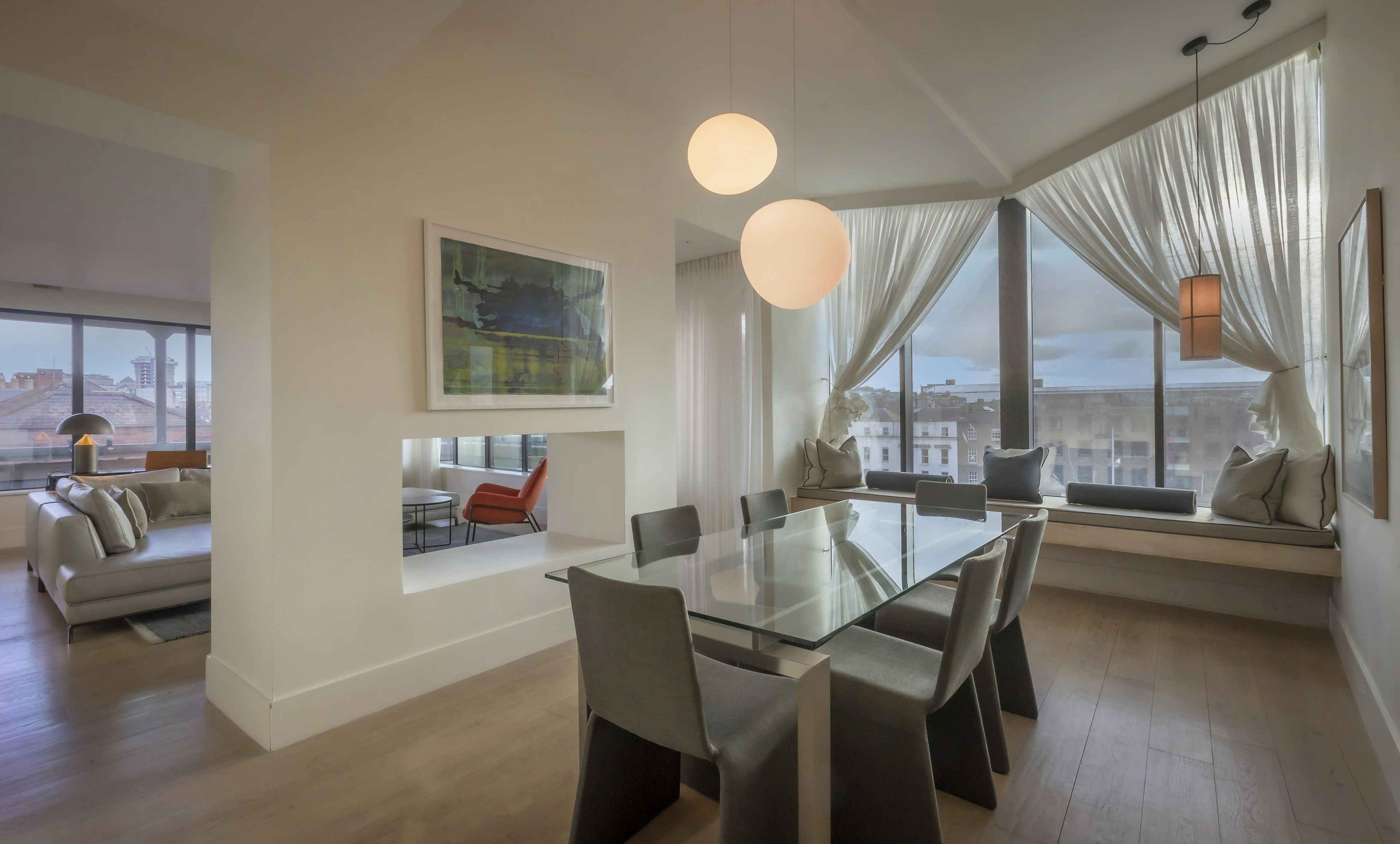 A modern living room with a glass dining table, comfortable seating, a large window with curtains, and a city view.