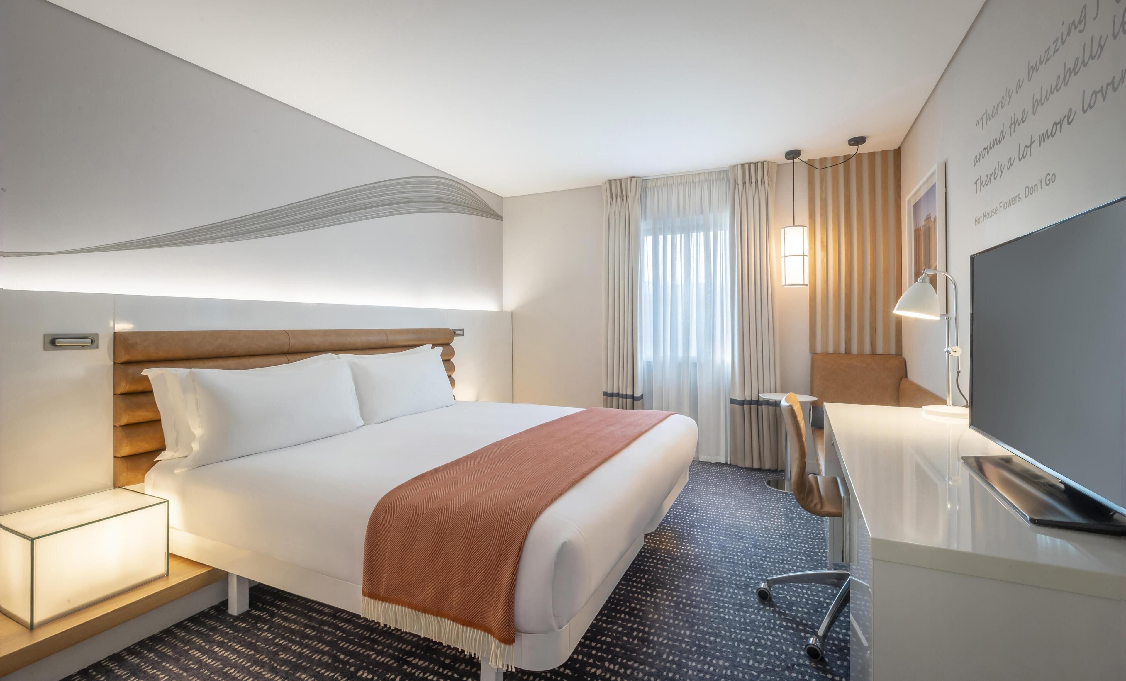 Una habitación de hotel con cama, mesita de noche, lámpara, televisor y una pared con una cita.