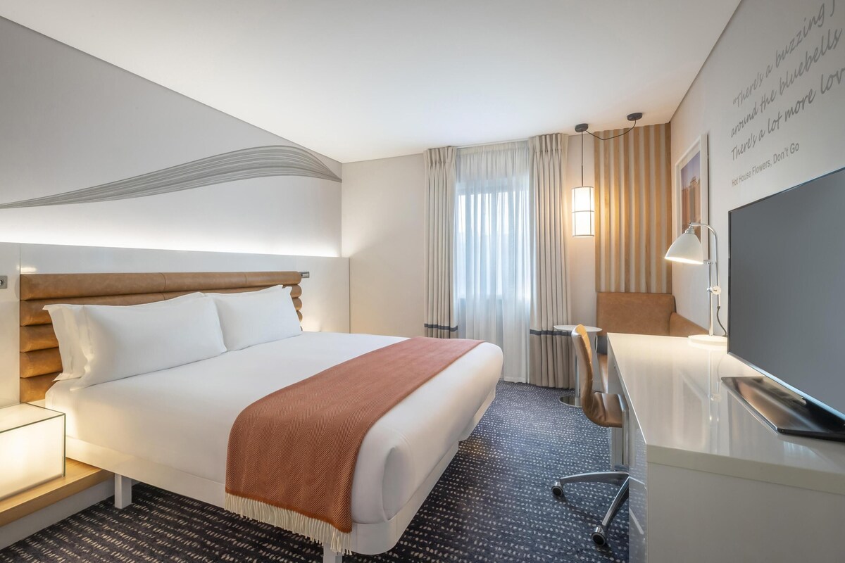 A hotel room with a bed, bedside table, lamp, TV, and a wall with a quote.