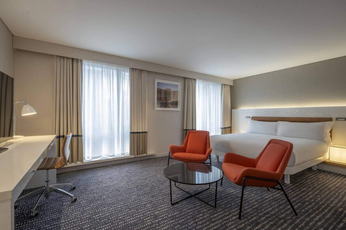 Quarto de hotel com uma cama, duas cadeiras laranja, uma mesa pequena, uma escrivaninha e um abajur.