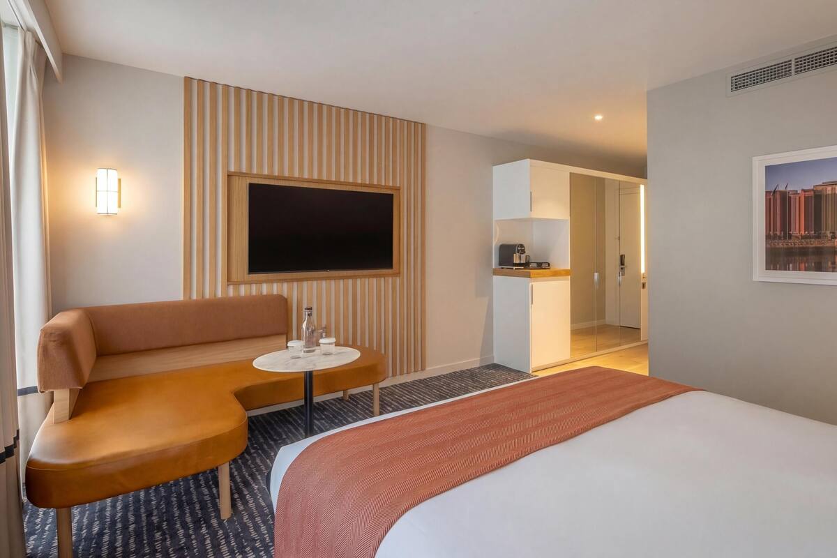 Quarto de hotel com cama, sofá, mesa pequena, TV, cozinha compacta e um quadro na parede.