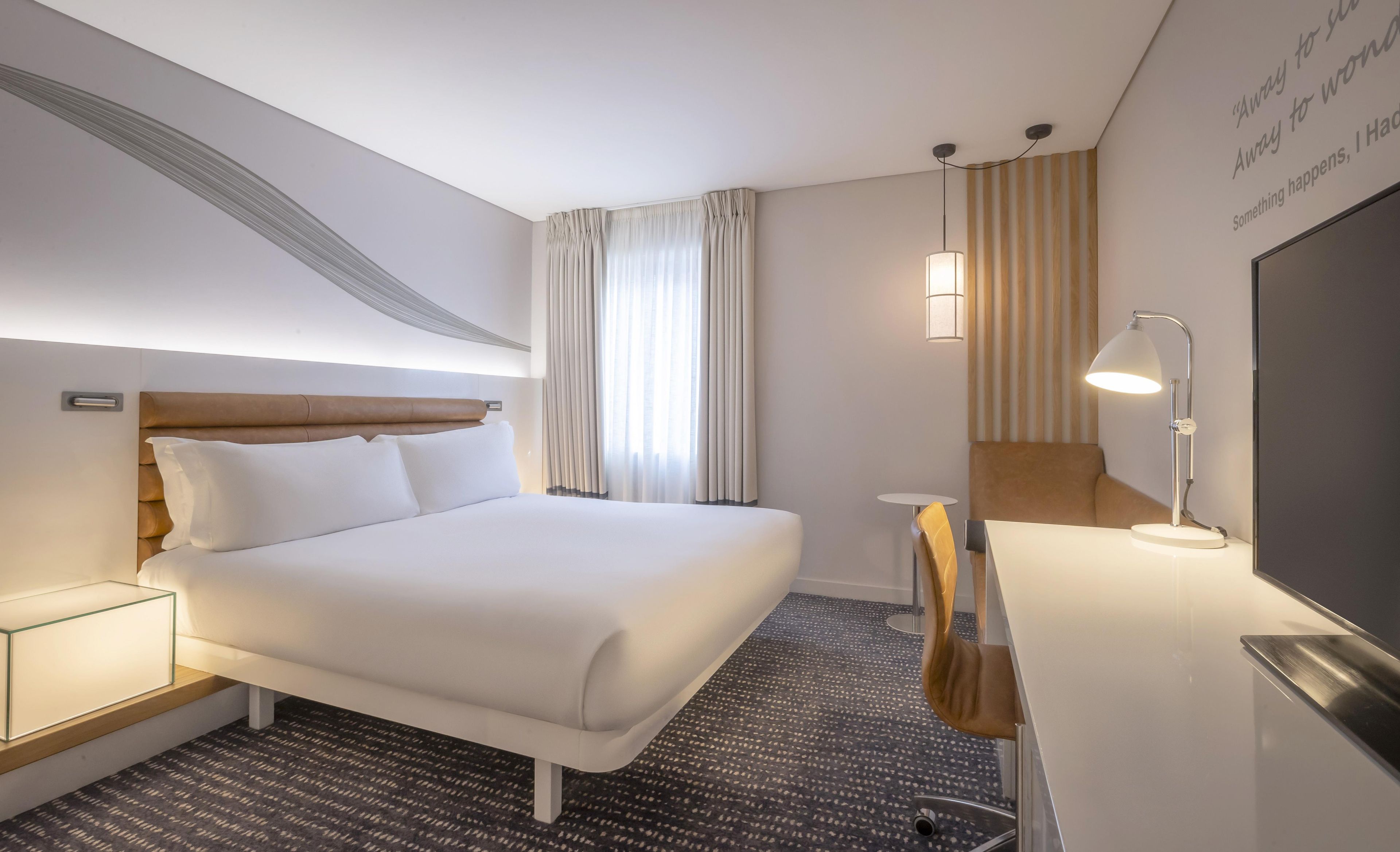 Ett hotellrum med en säng, ett nattduksbord, en lampa och ett skrivbord med en stol.