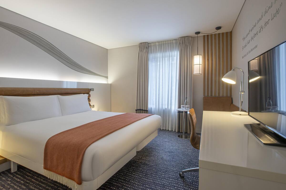Quarto de hotel com cama, mesa de cabeceira, luminária e espelho.