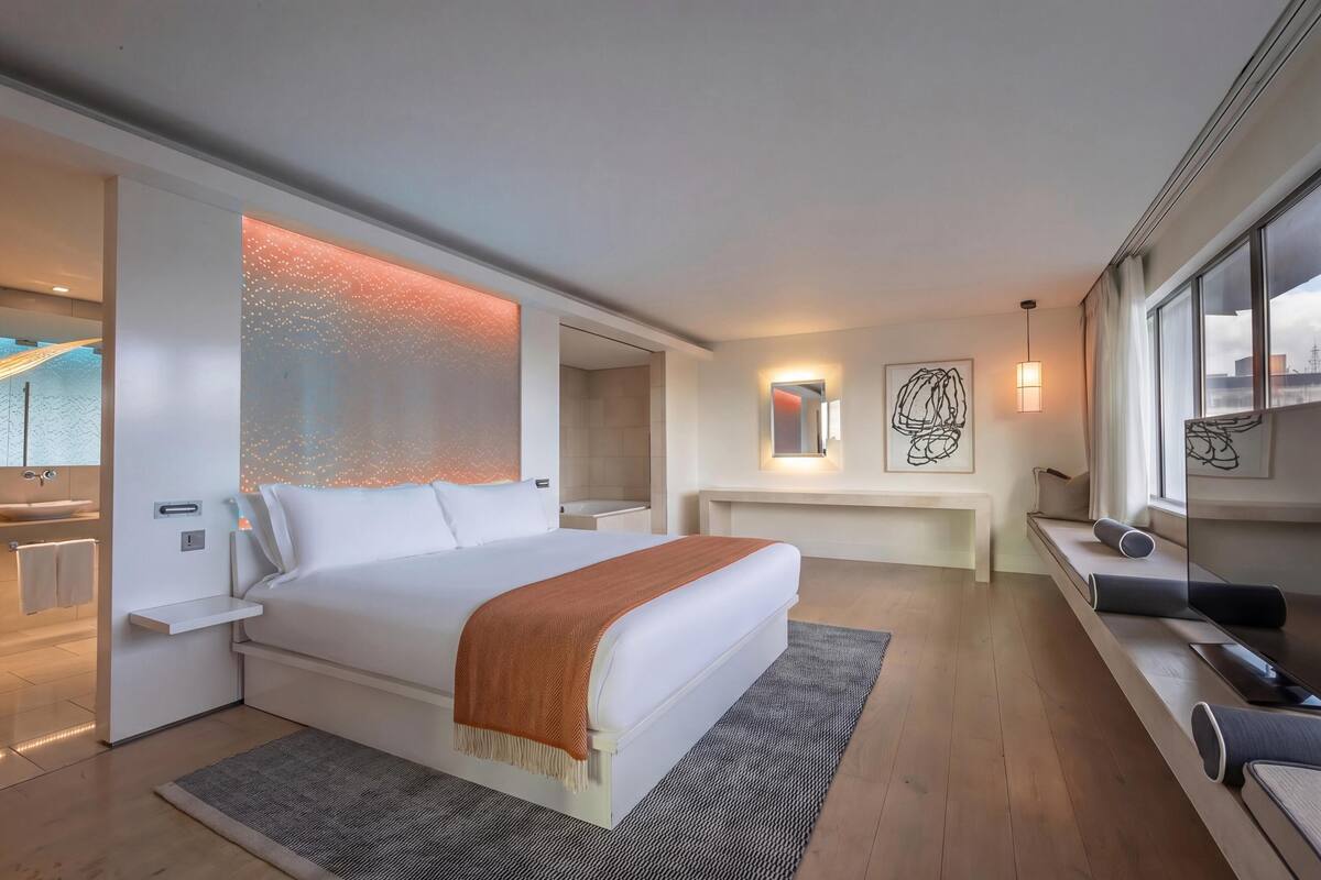 Quarto de hotel moderno com uma cama grande, mesas de cabeceira, um banco embutido com espaço de armazenamento, uma janela ampla e vista para a cidade.
