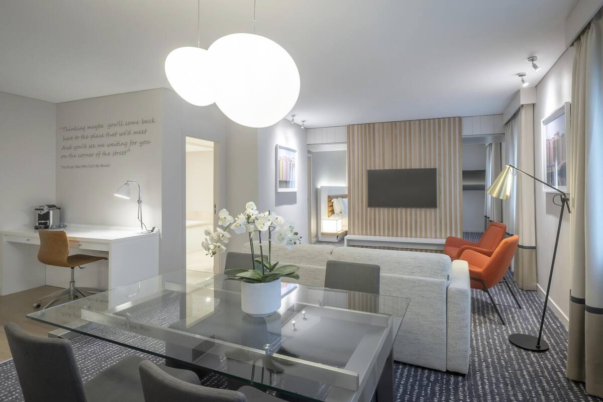 Quarto de hotel moderno com mesa de jantar de vidro, sofá, televisão e uma grande janela.
