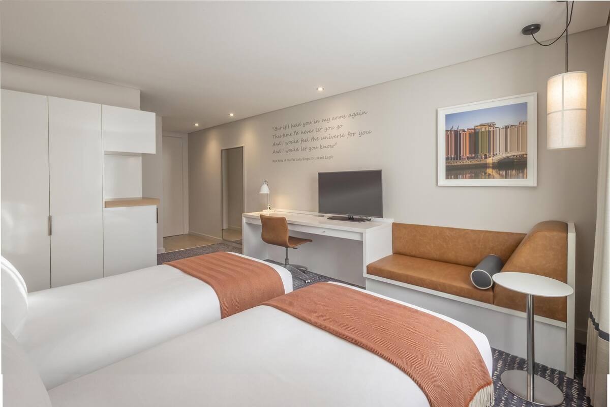 Quarto de hotel com duas camas, uma escrivaninha, uma cadeira, um sofá, uma televisão e uma parede com uma citação.