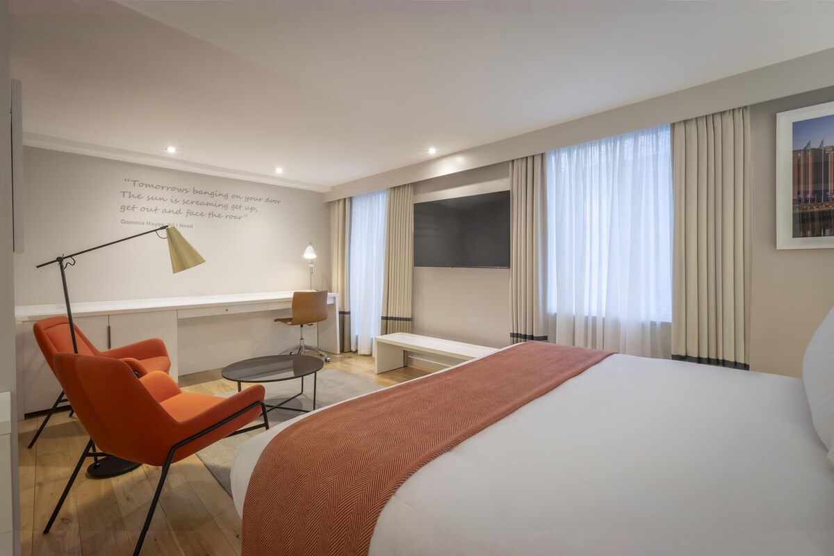 Ein Hotelzimmer mit einem Bett, einem Schreibtisch, einem Stuhl, einer Lampe und einem großen Fenster mit Vorhängen.