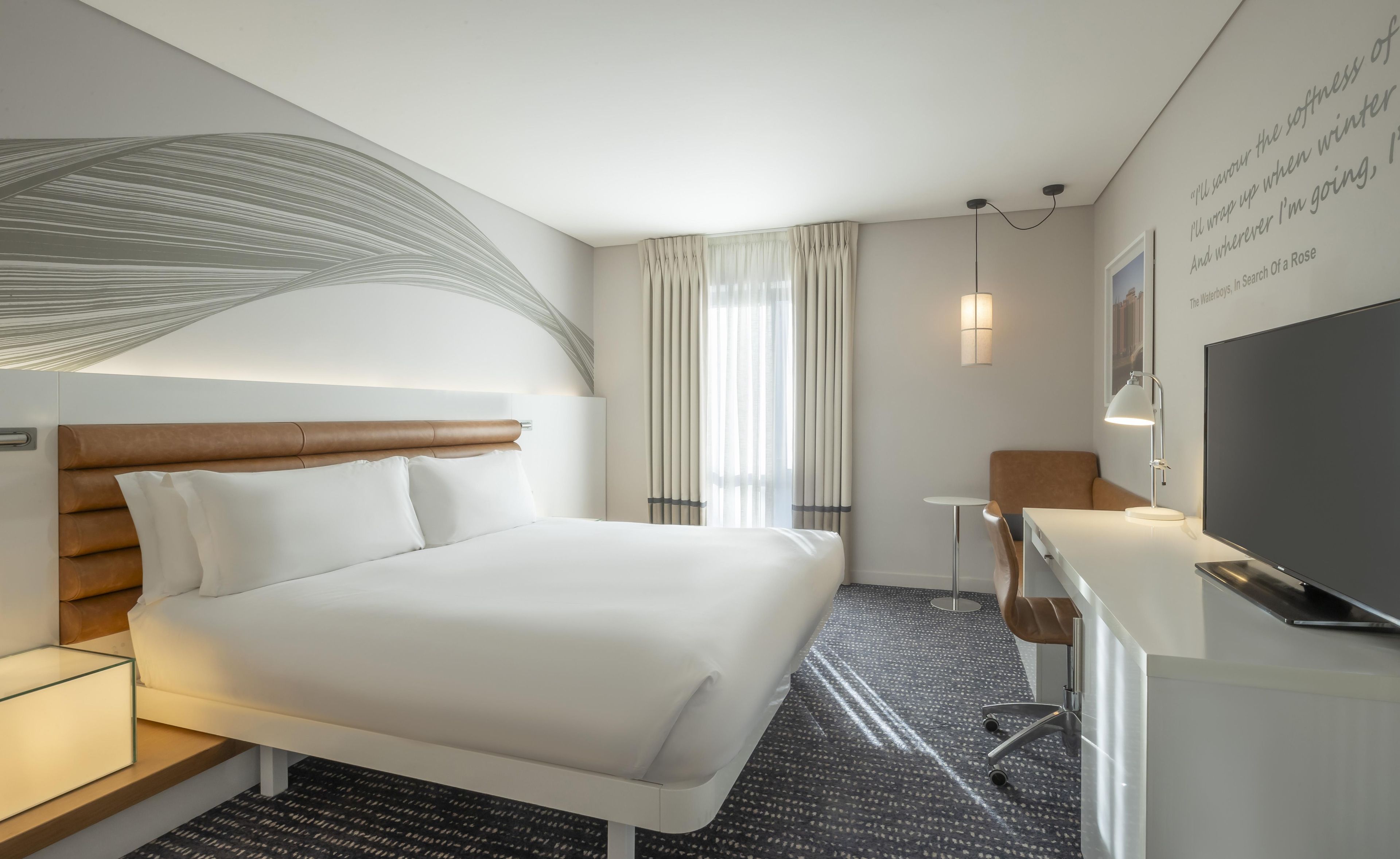 Habitación de hotel moderna con una cama grande, un escritorio, una silla, un televisor de pantalla plana y una lámpara de pared.
