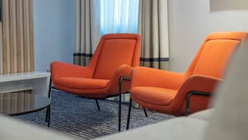 Ett hotellrum med två orangefärgade fåtöljer, ett litet bord och ett fönster med gardiner.