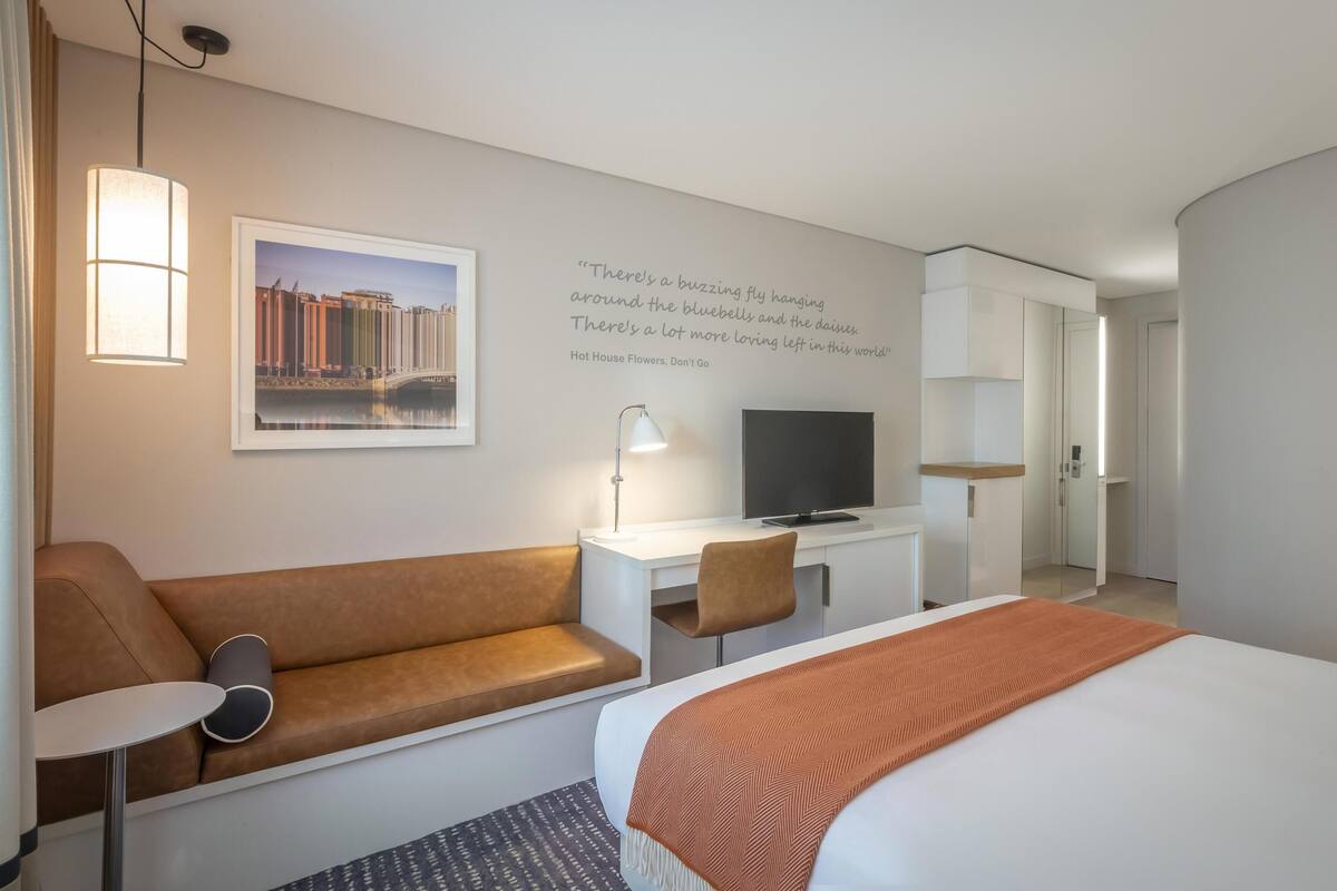 Quarto de hotel com cama, sofá, escrivaninha com televisão, luminária e parede com uma frase.