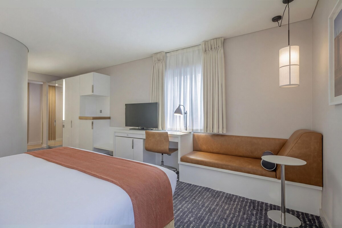 Ein Hotelzimmer mit einem Bett, einem Sofa, einem Schreibtisch mit Fernseher, einer Lampe und einem Fenster mit Vorhängen.