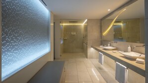 A modern bathroom with a large mirror, two sinks, a shower, and a bench.