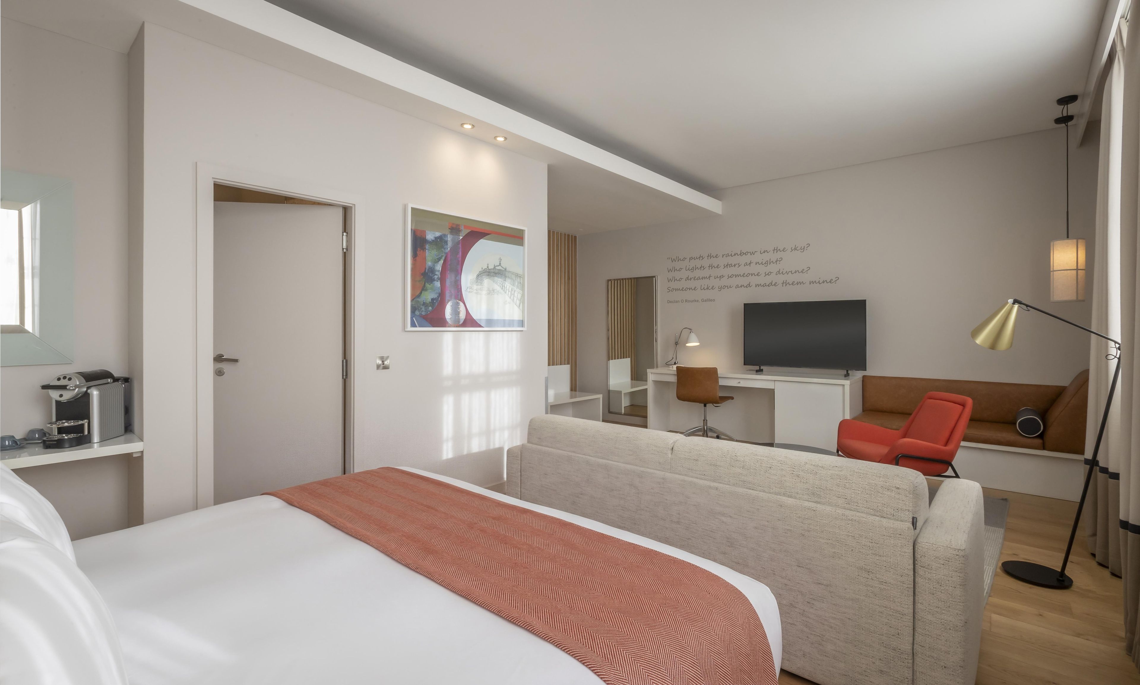 Habitación de hotel moderna con cama, escritorio, silla, televisor y lámpara de noche.