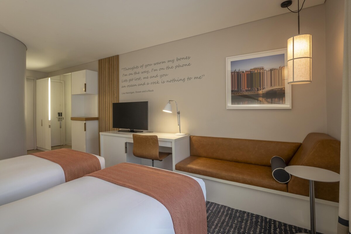 Ein Hotelzimmer mit zwei Betten, einem Schreibtisch mit Fernseher, einer Couch, einer Lampe und einem gerahmten Bild an der Wand.