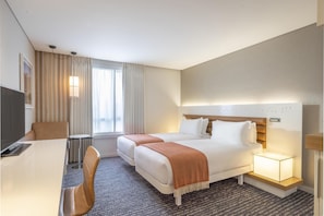 A hotel room with a bed, bedside table, lamp, TV, and a desk with a chair.