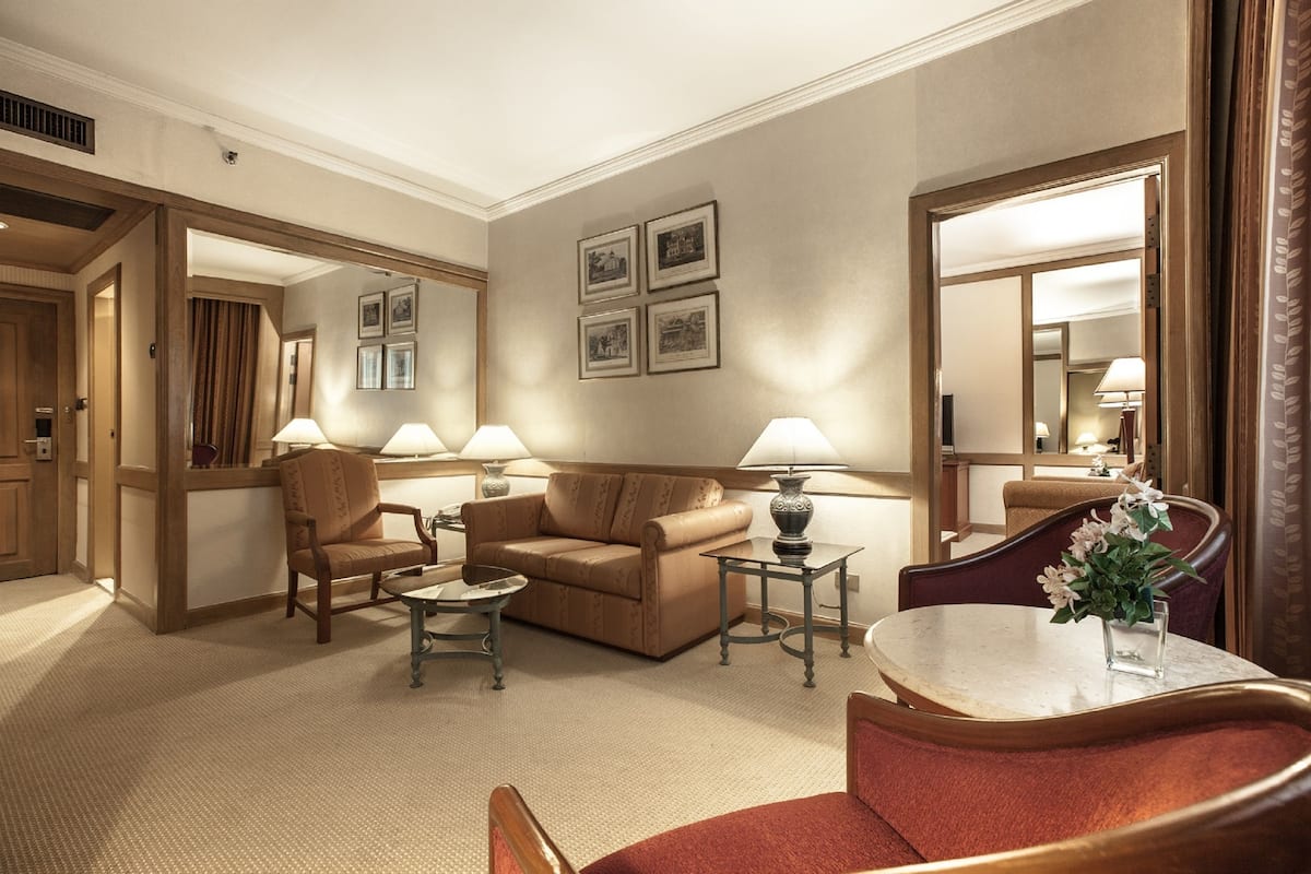 A hotel room with a living area featuring a sofa, armchairs, a coffee table, and a side table with lamps. There are framed pictures on the wall and a large mirror.