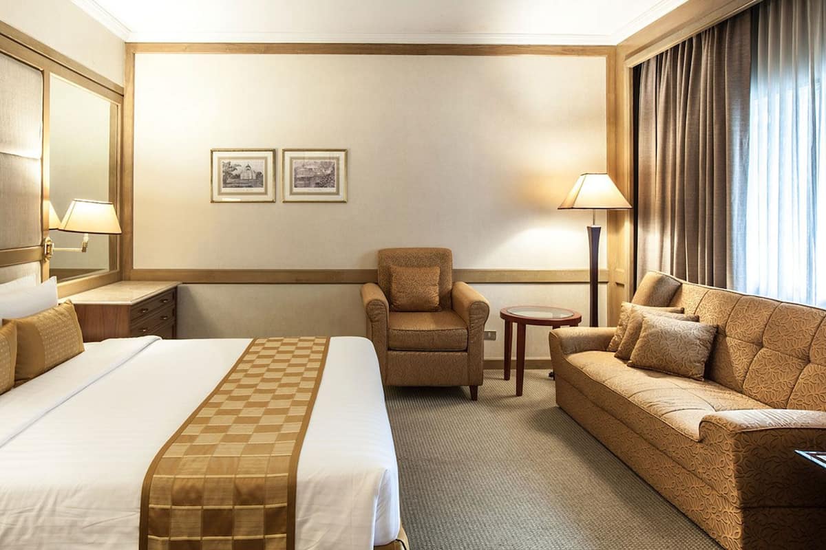A hotel room with a large bed, a sofa, a chair, and a small table. There are two framed pictures on the wall and a lamp on the side table.