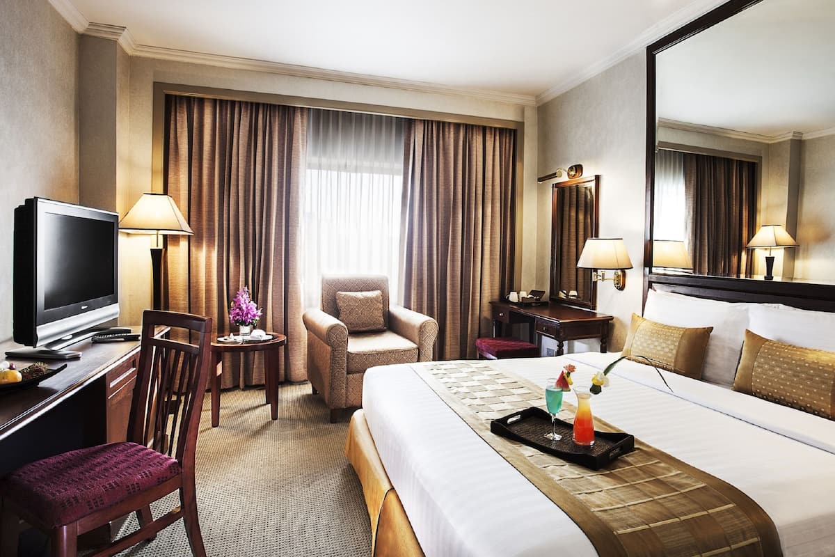 A hotel room with a bed, a desk with a television, a chair, a small table with a vase of flowers, and a large mirror on the wall.
