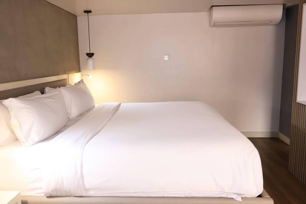 A neatly made bed with white linens, a hanging pendant light, and an air conditioning unit mounted on the wall.
