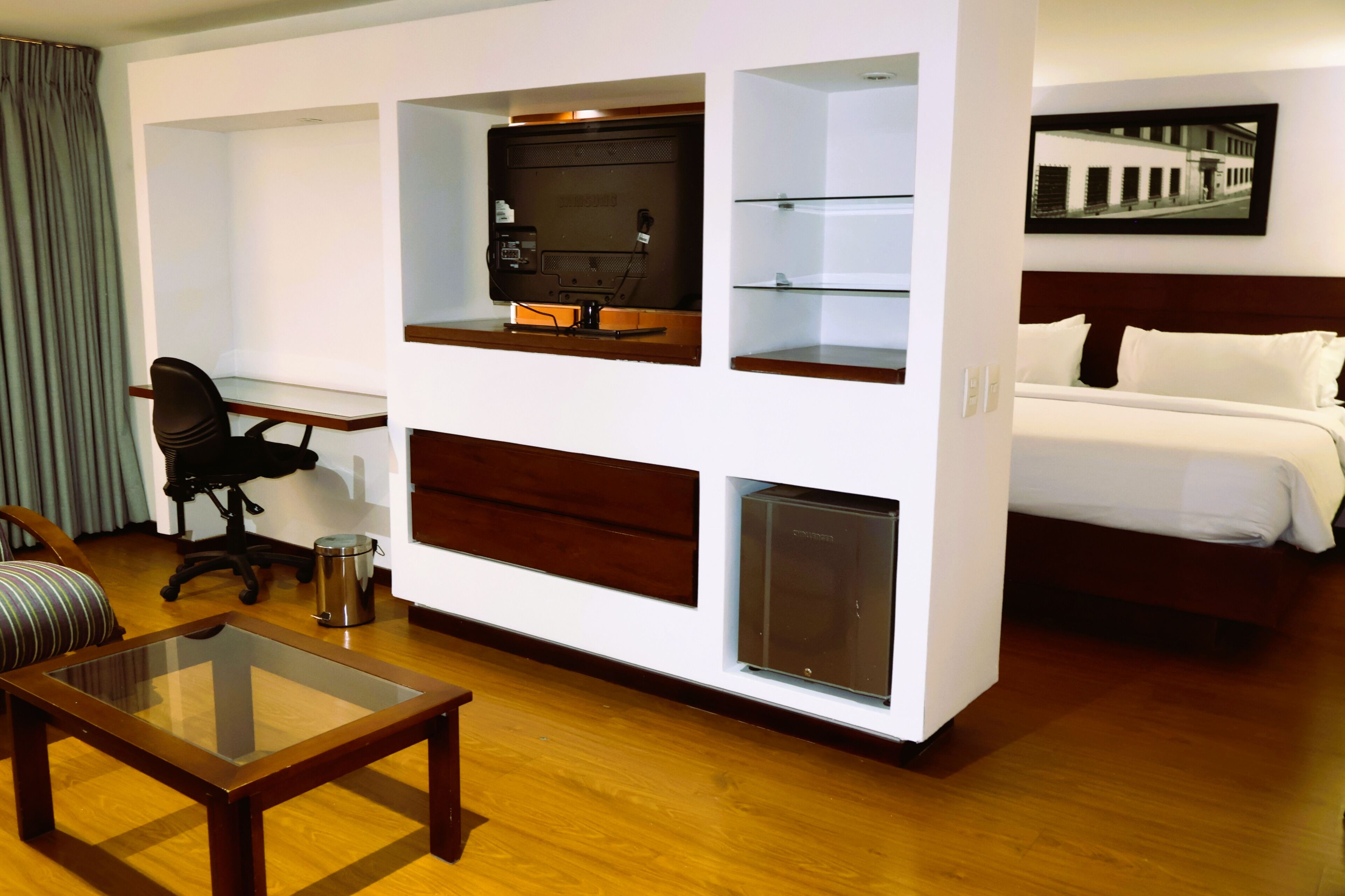 A hotel room with a bed, a desk with a computer, a glass coffee table, and a chair.