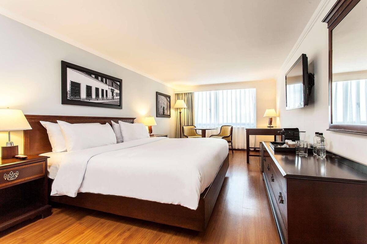 A hotel room with a large bed, bedside tables, a desk, a chair, a TV, and a window with curtains.