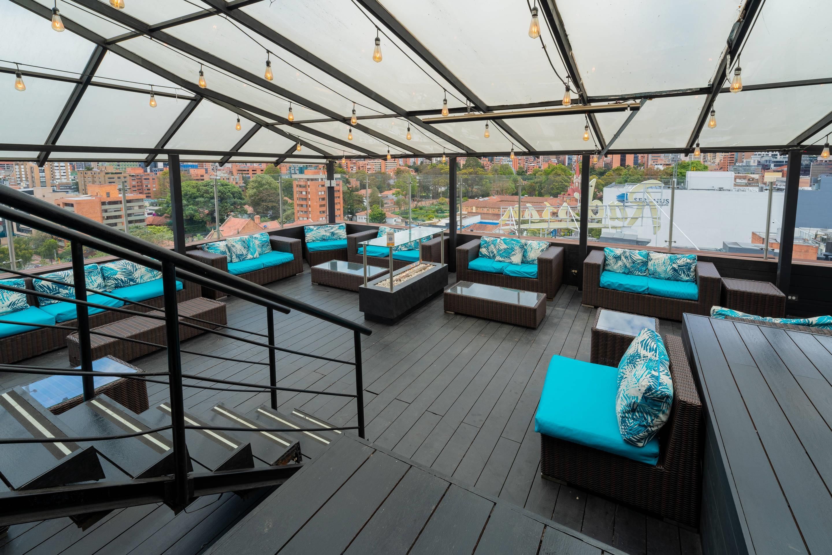 A rooftop lounge with wicker furniture and teal cushions, overlooking a cityscape.