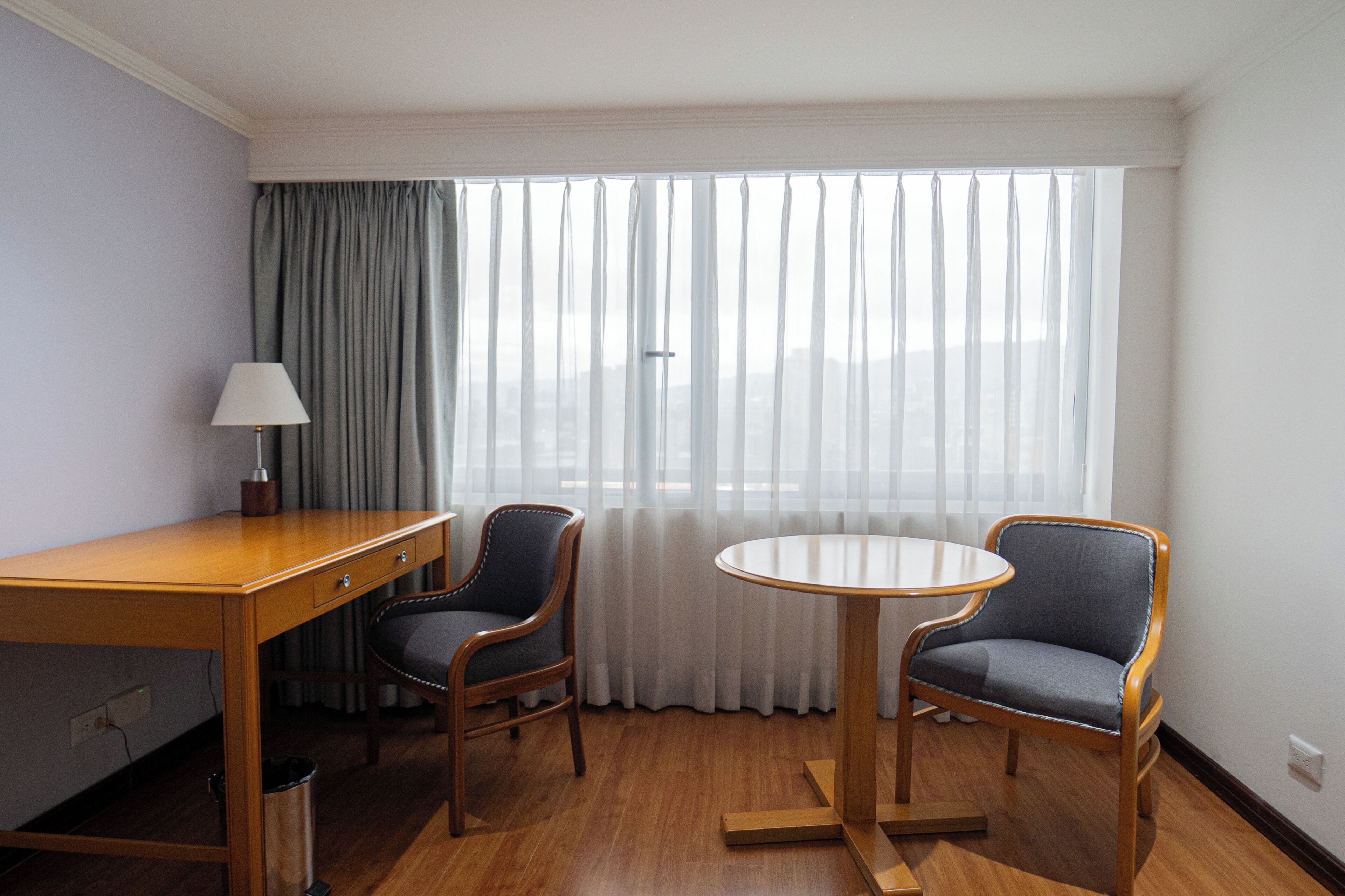 Quarto de hotel com secretária de madeira, duas cadeiras, uma mesinha, luminária e uma janela grande com cortinas.
