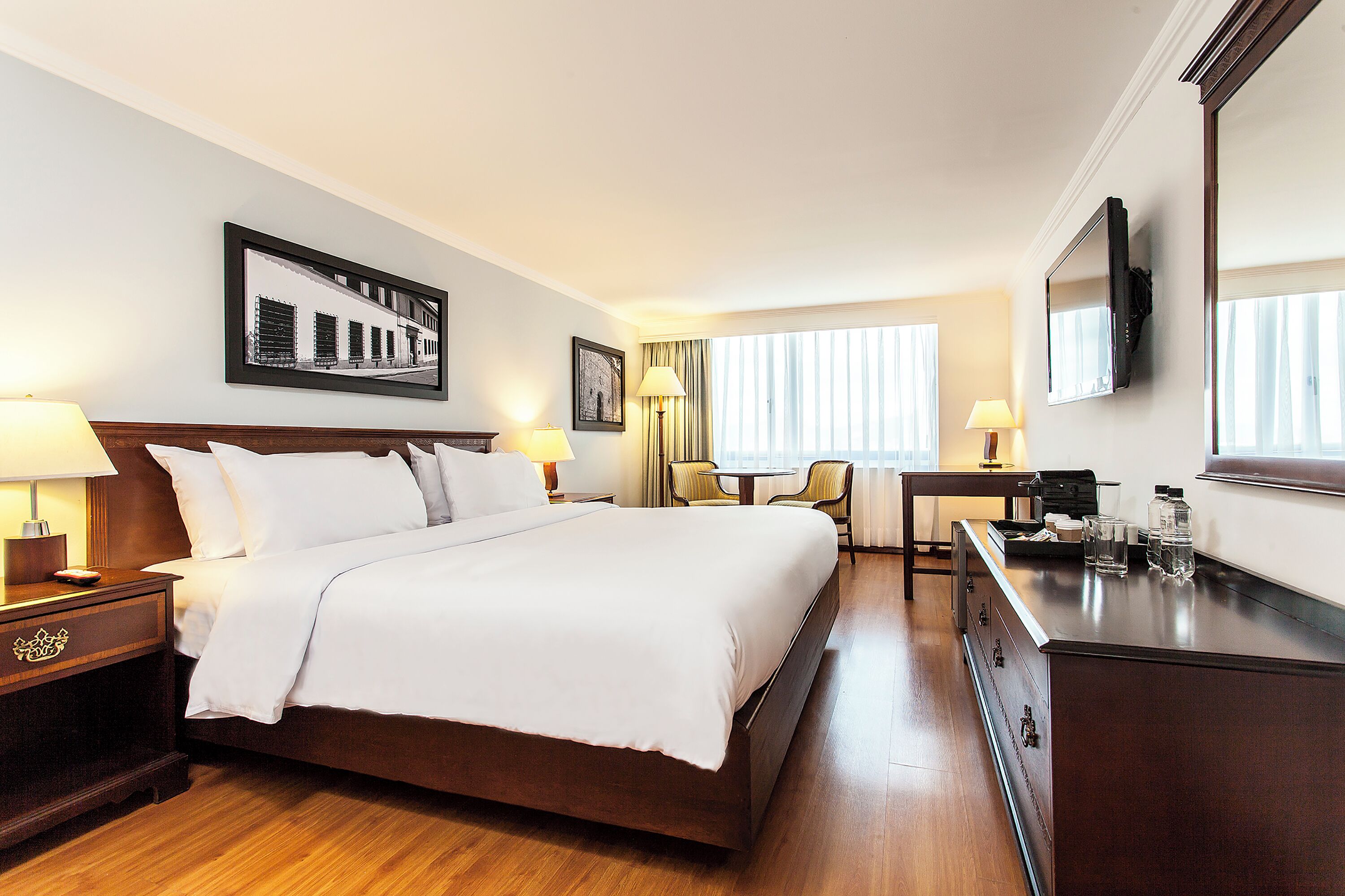 A hotel room with a bed, bedside tables, a desk, a chair, a TV, and a window with curtains.