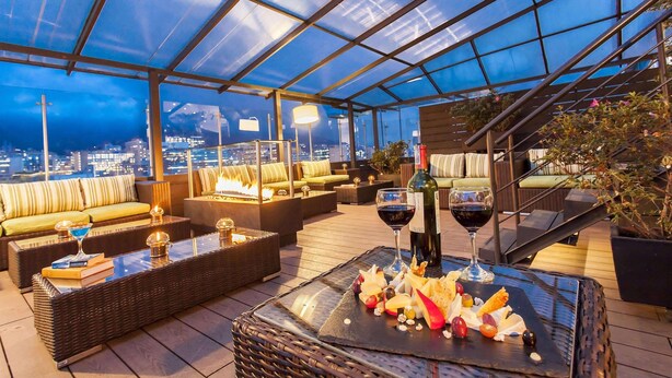 A rooftop terrace with wicker furniture, a glass table with food and wine, and a city view. - Radisson Bogota Metrotel (Bogotá)