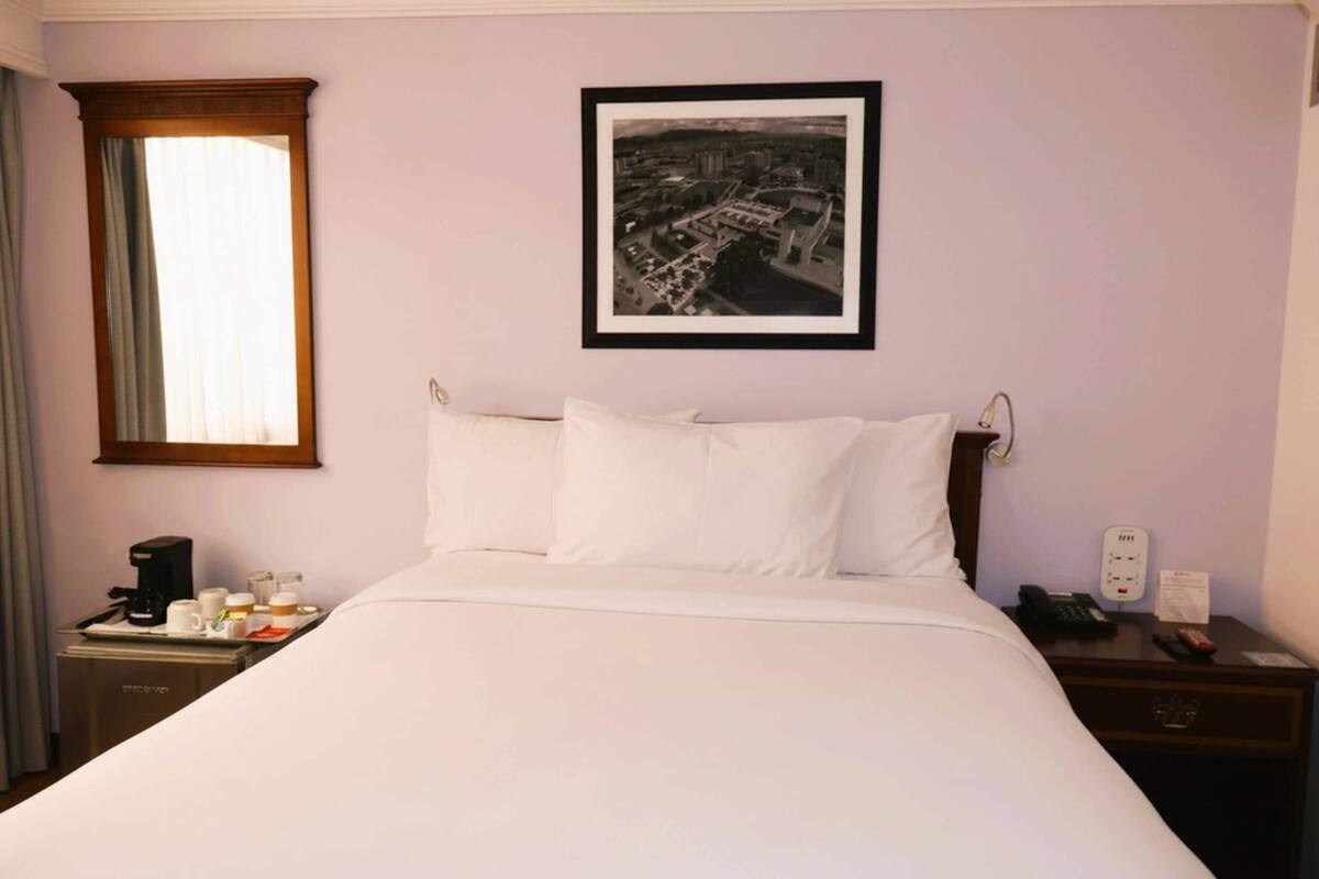 A hotel room with a bed, a nightstand with a phone and a lamp, a window with curtains, and a framed picture on the wall.