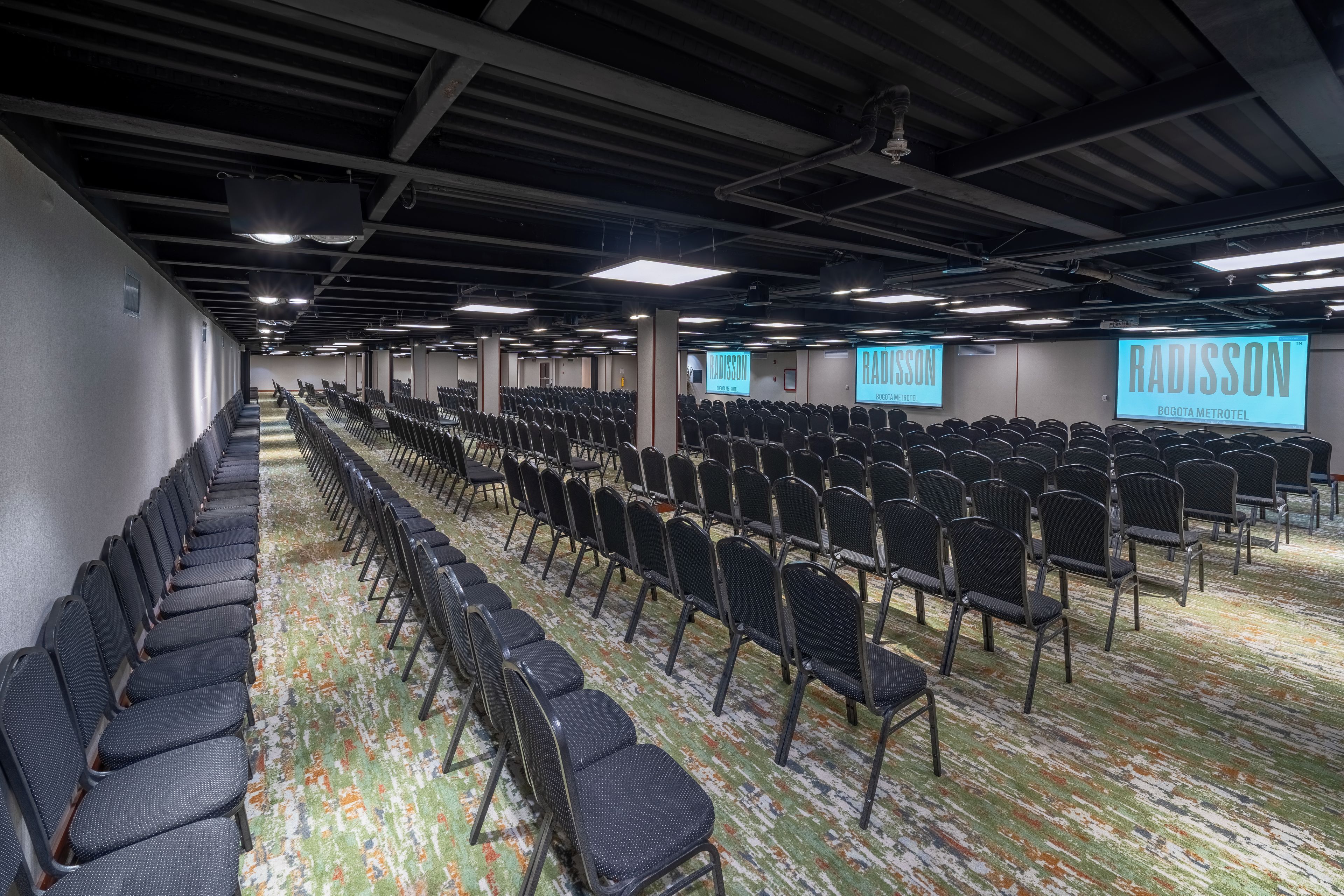Un salón de conferencias con filas de sillas, una pantalla de proyector y piso alfombrado.