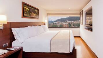 Un dormitorio de hotel con una cama grande, una mesita de noche con teléfono, un cuadro en la pared y vistas a la ciudad a través de la ventana.