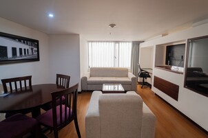 Un soggiorno con divano, tavolino, TV a parete, tavolo da pranzo con sedie e una cornice con un'immagine appesa al muro.