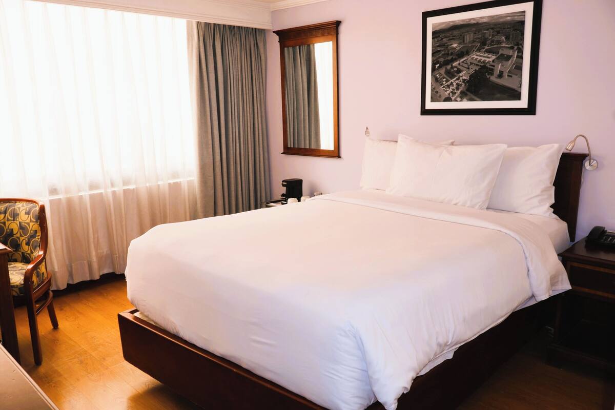A hotel room with a large bed, a chair, a nightstand, a framed picture on the wall, and a window with curtains.
