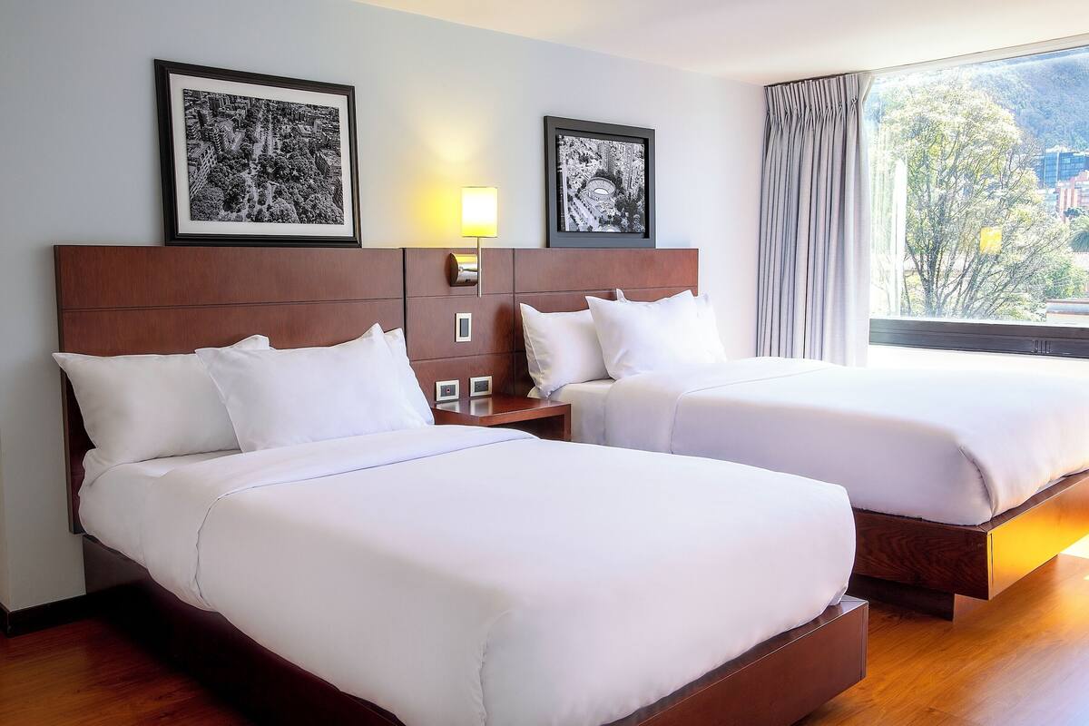 A hotel room with two beds, a nightstand, a lamp, framed pictures on the wall, and a window with a view of trees and buildings.