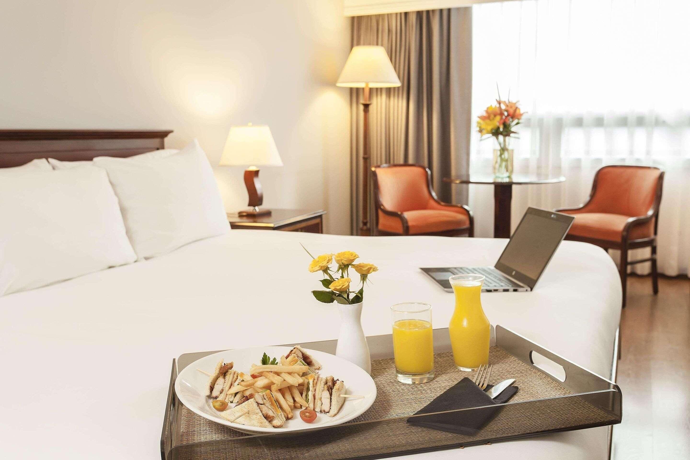 A hotel room with a bed, a laptop on the bed, a tray with food and drinks, and a vase with flowers.